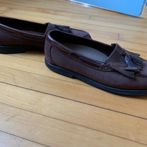 Peut inclure: Une paire de mocassins en cuir marron avec des glands. Les mocassins ont une tige en cuir marron fonc&eacute; et une semelle en caoutchouc noir.