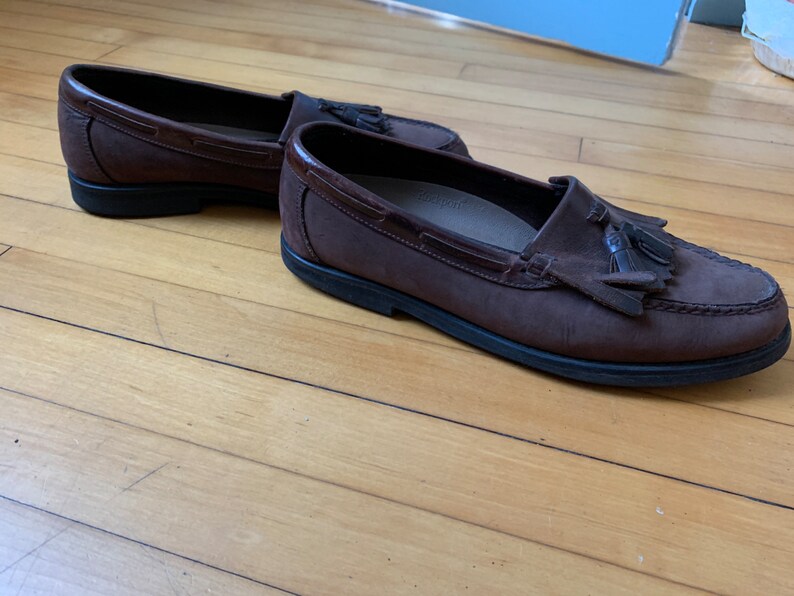Peut inclure: Une paire de mocassins en cuir marron avec des glands. Les mocassins ont une tige en cuir marron fonc&eacute; et une semelle en caoutchouc noir.