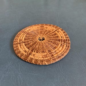 May include: A circular, flat woven coaster crafted from natural pine needles. The design features a radiating pattern of light tan and dark brown fibers coiled in concentric rings, with a small open center point and intricate stitching throughout.