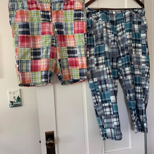 May include: Two pairs of patchwork plaid shorts and pants hanging on a white door. The shorts are a mix of red, pink, green, and blue squares. The pants are blue, white, and gray squares.