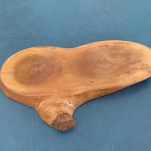 May include: A handcrafted, wooden serving tray with a unique, organic shape. The tray is made from a light brown wood, featuring visible grain patterns and a natural, unfinished look. The tray has a rounded, asymmetrical design with a small, protruding handle.