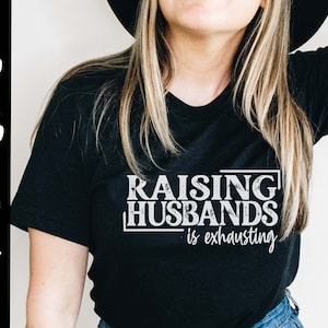 Puede incluir: Una camiseta negra con texto blanco que dice "Raising Husbands is exhausting".