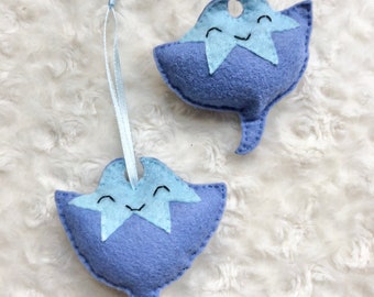 May include: Two blue felt ornaments shaped like stingrays with smiling faces and a light blue star on their bodies. One ornament has a white ribbon attached to it.