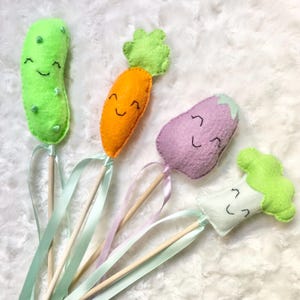 May include: Four felt vegetable wands: a green cucumber, orange carrot, purple eggplant, and white broccoli. Each has a smiling face and is attached to a wooden stick with a ribbon. The vegetables are handmade.