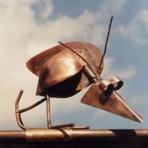 Könnte beinhalten: Eine Kupferskulptur eines Vogels mit langem Schnabel und großen Augen. Der Vogel sitzt auf einem braunen Metallrohr.