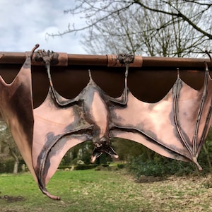 May include: A copper bat sculpture hanging from a gutter. The bat's wings are spread wide and its body is facing down. The sculpture is made of detailed copper with a patina finish.