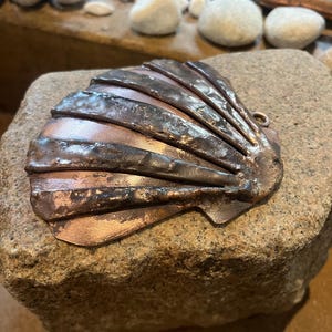 Könnte beinhalten: Ein metallisches Jakobsmuschel-Ornament mit kupferfarbenem Finish und einer texturierten, gehämmerten Oberfläche. Die Muschel weist ausgeprägte radiale Rippen und eine kleine Öse an der Oberkante zum Aufhängen auf.