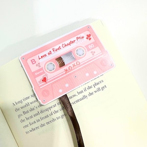 May include: A pink cassette tape bookmark with the text "Love at First Chapter Mix" and "XOXO". The bookmark is inserted into a book with printed text and a brown ribbon. The cassette tape design includes hearts and the number 60.
