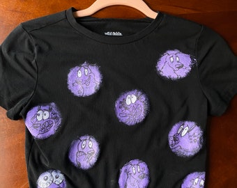 May include: Black cropped t-shirt with purple cartoon images of Courage the Cowardly Dog. The shirt has the text "Wild Fable" on the neck.