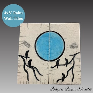 May include: Two 4x8 inch Raku wall tiles with a white background and black and blue designs. The design features a large blue circle and black silhouettes of birds on branches.