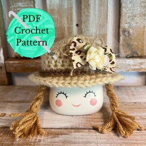 May include: A crochet pattern for a cute, brown and tan hat with a leopard print bow and a white flower. The hat is sitting on a white mug with a smiling face and brown yarn pigtails.