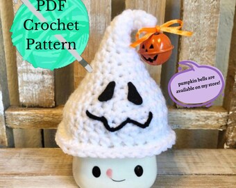 May include: A white crocheted ghost with a black embroidered face and a pumpkin bell on top. The ghost is sitting on a white ceramic base with a smiling face. The text "PDF Crochet Pattern" is in the top left corner of the image. The text "pumpkin bells are available on my store!" is in a purple speech bubble in the bottom right corner of the image.