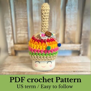 Puede incluir: Un patrón de crochet para un calientavasos colorido a rayas con un botón marrón y un pequeño lazo. El calientavasos está hecho con un hilo beige y tiene una carita feliz en la parte inferior. Patrón de crochet PDF, términos estadounidenses, fácil de seguir.