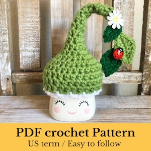 Puede incluir: Un patrón de crochet para un gorro de gnomo verde con una flor blanca y una mariquita en una hoja verde. El gorro está sentado en una taza blanca con una cara sonriente. El texto "PDF crochet Pattern US term / Easy to follow" está en la parte inferior de la imagen.