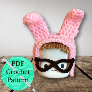 May include: A pink crocheted bunny hat with brown crocheted glasses. The hat is on a white mug with a brown mustache and a smiling face. The text "PDF Crochet Pattern" is on a green ball of yarn with a crochet hook.