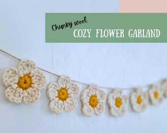 Op de afbeelding: Een gezellige bloemenslinger van dikke wol. De slinger heeft meerdere gehaakte bloemen met crèmekleurige bloemblaadjes en gele centra, geregen aan een dun, natuurlijk gekleurd koord. De tekst "Chunky wool COZY FLOWER GARLAND" staat erboven.