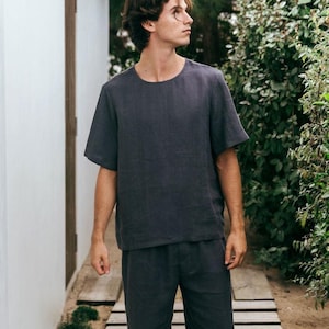 Peut inclure: Un ensemble en lin gris foncé composé d'une chemise à manches courtes et d'un pantalon assorti. La tenue a une coupe décontractée et est fabriquée dans un tissu léger. Le modèle se tient sur une passerelle en bois à l'extérieur.