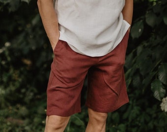Op de afbeelding: Een man in een wit linnen shirt en een donkerrode broek staat voor een groene, bladerrijke achtergrond. Hij draagt witte sokken en zwarte sneakers.