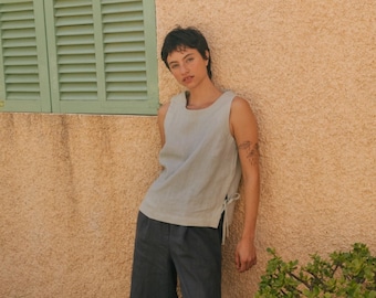 May include: A person wearing a light blue sleeveless top with a side tie detail and dark blue wide-leg pants. They are standing in front of a yellow wall with a green window shutter.