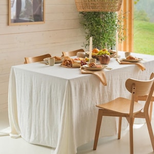 May include: A dining table set for a meal, featuring a white linen tablecloth, wooden chairs, and a woven pendant light. The table is set with plates of food, fruit, and croissants. A framed picture hangs on the wall.