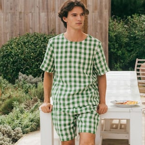 May include: A person wearing a green and white checkered short-sleeved shirt and matching shorts. The outfit is made of a lightweight fabric. The person is standing near a white table outdoors.