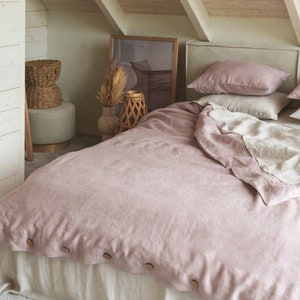 Peut inclure: Une housse de couette en lin rose avec des boutons en bois sur un lit avec une tête de lit en lin brun clair et un couvre-lit en lin beige. Le lit est dans une pièce avec un plafond en bois et un panier en osier sur un tabouret.