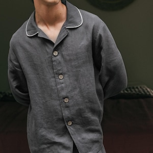 May include: Charcoal gray pajama set with white piping on the collar and cuffs. The button-down shirt has three buttons and is paired with matching pants. The fabric appears to be linen or a similar lightweight material.