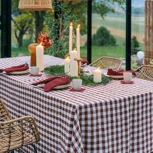 May include: A red and white gingham tablecloth is set on a table in a greenhouse. The table is decorated with candles, greenery, and wicker chairs.