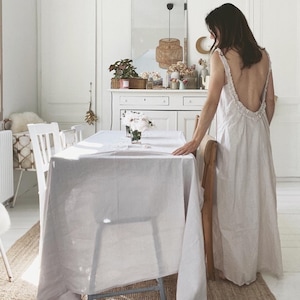 Puede incluir: Una mesa de comedor blanca con un mantel blanco y sillas. Una mujer con un vestido blanco está de pie junto a la mesa. La habitación tiene paredes blancas y una lámpara de mimbre colgando del techo.