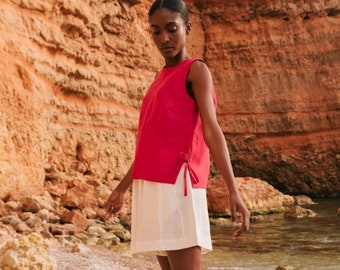 May include: A woman wearing a bright pink sleeveless top with a white linen skirt and pink sandals stands on a rocky beach.