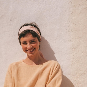 May include: A woman wearing a light yellow linen dress with a pink headband. She is sitting on a stone step with a white wall behind her.