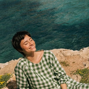 May include: A woman wearing a green and white gingham dress sits on a rocky cliff overlooking the sea. She is smiling and has her eyes closed.