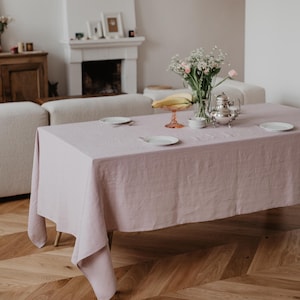 May include: A light pink linen tablecloth is laid on a wooden table with a herringbone pattern. The table is set with two white plates, a glass bowl with fruit, a silver teapot, and a vase of white flowers.