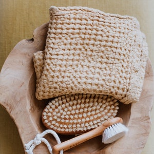 Op de afbeelding: Een houten kom met een beige wafel geweven handdoek, een houten borstel met natuurlijke borstels en een kleinere witte borstel met een houten handvat.