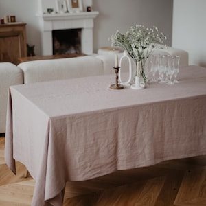 May include: A light pink linen tablecloth with a white ceramic vase filled with white flowers, two candlesticks, and a set of clear glass wine glasses on a wooden floor.