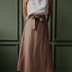 Könnte beinhalten: Eine Frau trägt ein weißes Tanktop und einen langen, braunen Leinenrock, der mit einem braunen Band in der Taille gebunden ist. Sie steht auf einem grauen Fliesenboden.