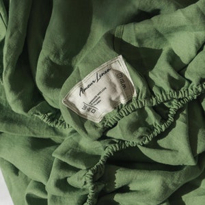 Puede incluir: Sábana bajera de lino verde con una etiqueta blanca que dice "Amour Linen" e incluye instrucciones de cuidado. La etiqueta está unida a la sábana con un hilo blanco.