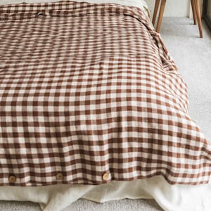 Pode incluir: Uma capa de edredom em gingham marrom e branco com botões de madeira. A capa de edredom está em uma cama com fronhas combinando.