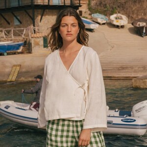 K&ouml;nnte beinhalten: Eine Frau in einem wei&szlig;en Leinen-Wickeltop und einer weiten gr&uuml;nen und wei&szlig;en Gingham-Hose steht auf einem Steg am Wasser. Sie blickt in die Kamera. Im Hintergrund sind Boote und ein kleines Dorf zu sehen.