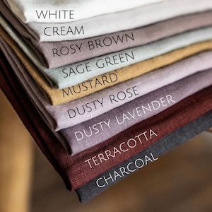 Pode incluir: Uma pilha de amostras de tecido em várias cores, incluindo branco, creme, marrom rosado, verde sálvia, mostarda, rosa empoeirado, lavanda empoeirada, terracota e carvão.