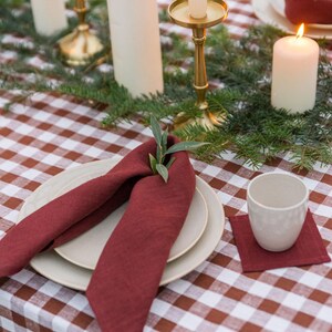 May include: A red linen napkin folded into a decorative shape with a sprig of greenery on top. The napkin is placed on a white and brown gingham tablecloth with two white plates underneath. There are candles and candle holders on the table.