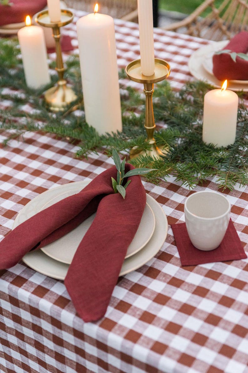 May include: A red linen napkin folded into a decorative shape with a sprig of greenery on top. The napkin is placed on a white and brown gingham tablecloth with two white plates underneath. There are candles and candle holders on the table.
