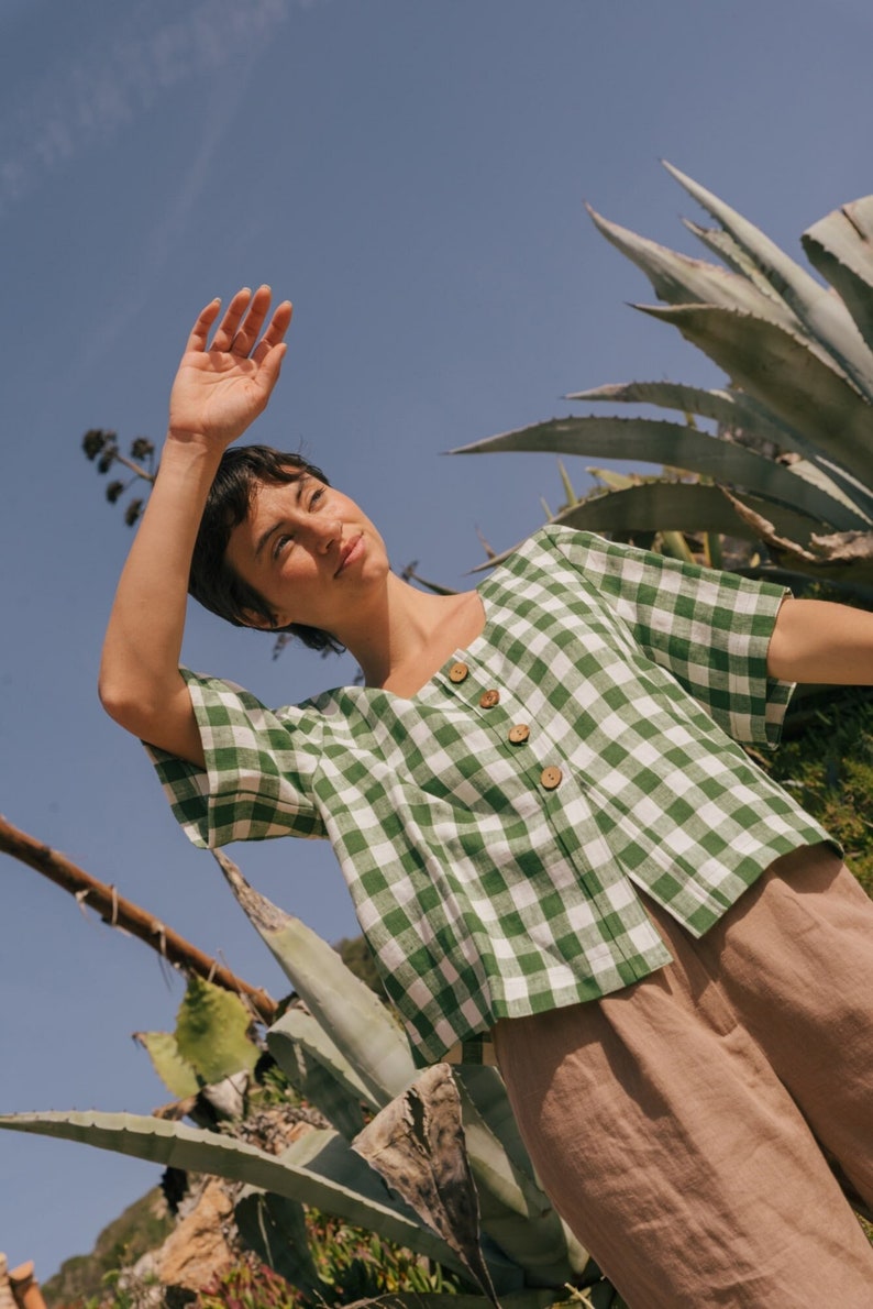 May include: A woman wearing a green and white gingham button-up shirt with short sleeves and brown pants. She is standing in front of a large green plant.