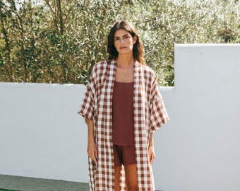 Op de afbeelding: Een bruine en witte gingham-badjas met een lange, vloeiende silhouet. De badjas heeft een ontspannen pasvorm en wordt gedragen door iemand die op een houten dek bij een zwembad staat.