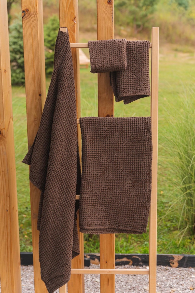 Op de afbeelding: Vier bruine wafeldoek handdoeken hangen aan een houten rek. De handdoeken zijn van verschillende grootte en zijn allemaal gemaakt van een zacht, absorberend materiaal.
