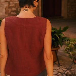 May include: A woman wearing a sleeveless, rust-colored linen top with a scoop neck and a dark gray skirt. She has short, dark hair and a tattoo on the back of her neck.