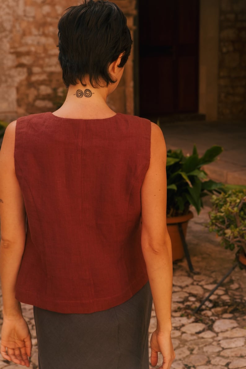 May include: A woman wearing a sleeveless, rust-colored linen top with a scoop neck and a dark gray skirt. She has short, dark hair and a tattoo on the back of her neck.