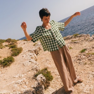 May include: A woman wearing a green and white gingham button-up shirt with short sleeves and brown wide-leg pants. She is standing on a rocky cliff overlooking the ocean.
