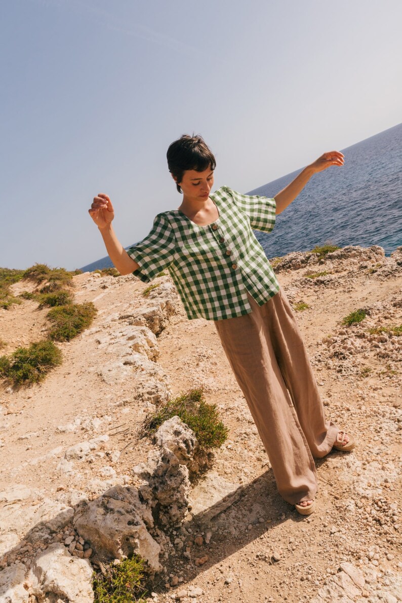 May include: A woman wearing a green and white gingham button-up shirt with short sleeves and brown wide-leg pants. She is standing on a rocky cliff overlooking the ocean.