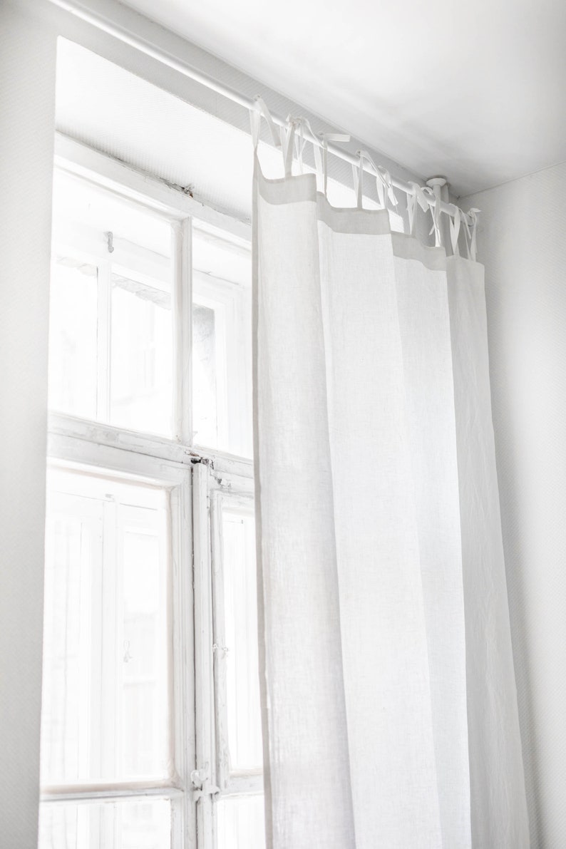 A white linen curtain with tie-tops hung on a tension rod inside a white window frame. A white linen curtain with tie-tops hung on a tension rod inside a white window frame.